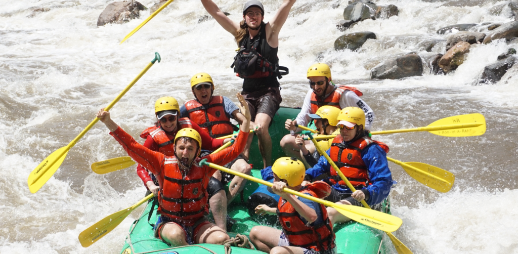 People in a green raft and yellow helmets hold their arms up after going through a rapid