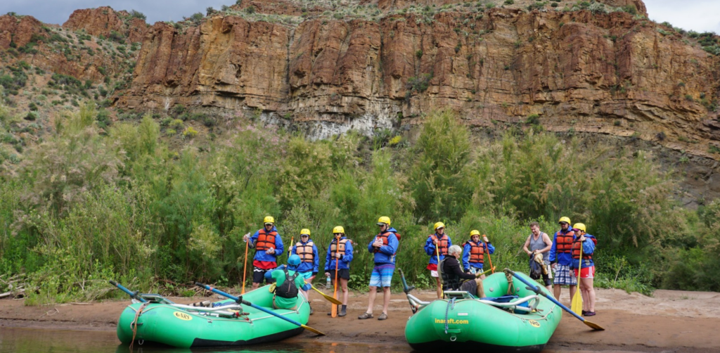 People stand next to green rafts and a red canyon wall on the Salt River in Arizona