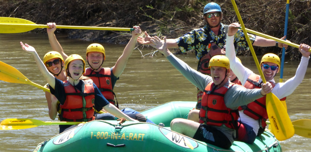 People in a green raft hold their yellow paddles up and smile on the salt river in Arizona